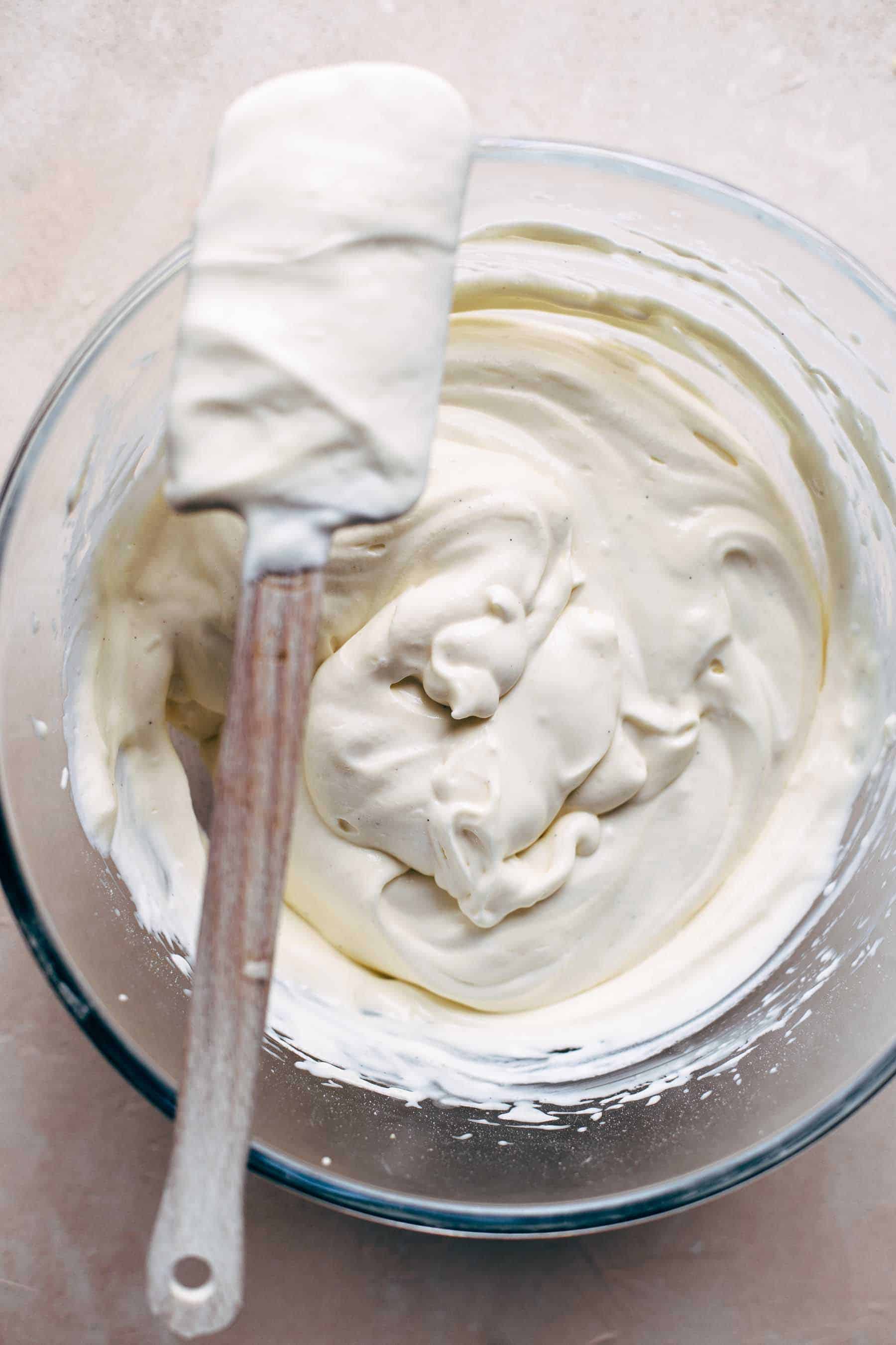 A bowl of whipped mascarpone cream with a spatula covered in cream on top of the bowl