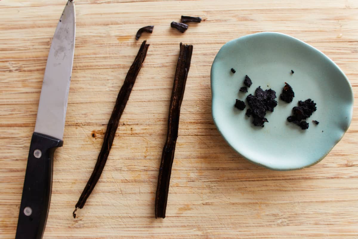 two split vanilla pods with seeds removed on a wooden board with a knife