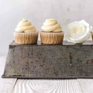 two cupcakes with frosting sitting on an inverted loaf pan with a white rose next to it