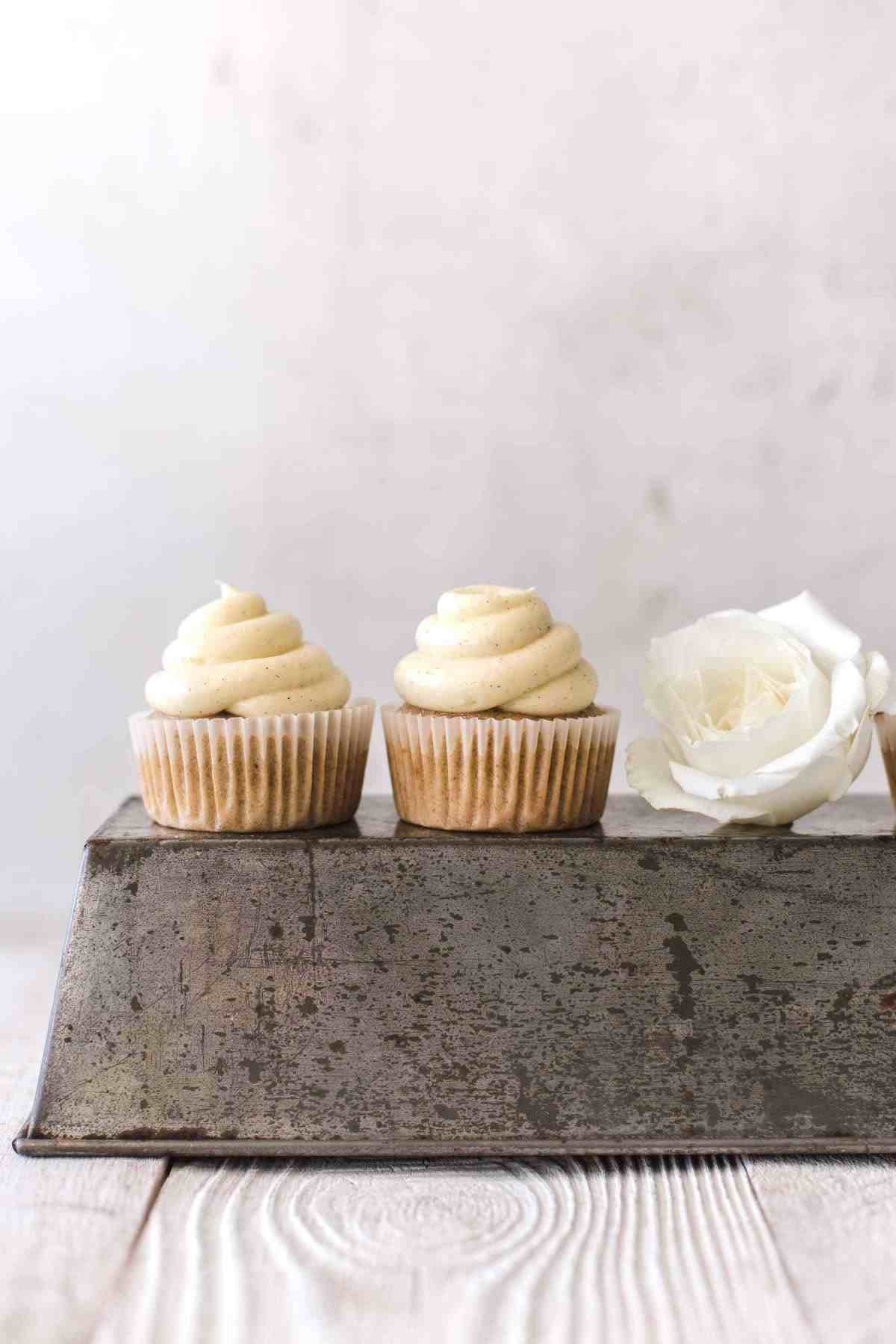 two cupcakes with frosting sitting on an inverted loaf pan with a white rose next to it