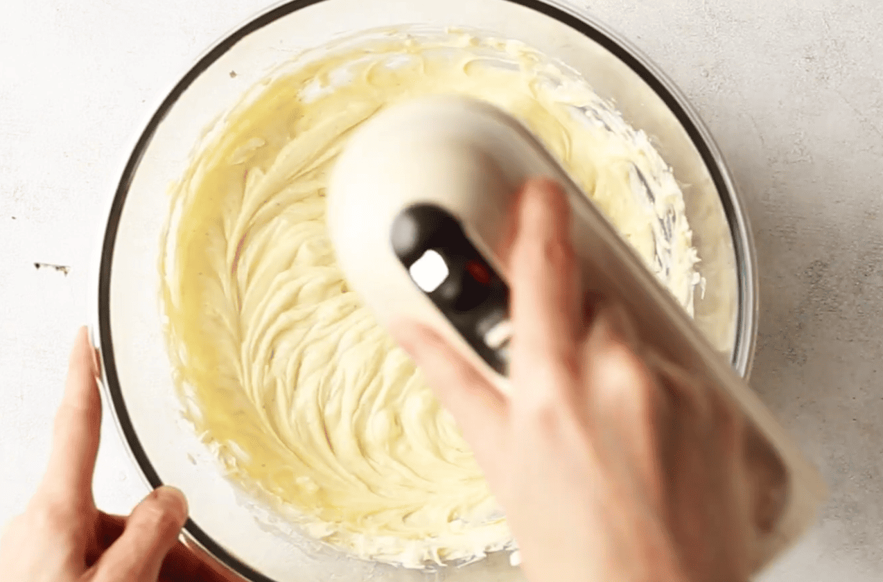 beating cream cheese in a glass bowl using hand mixer