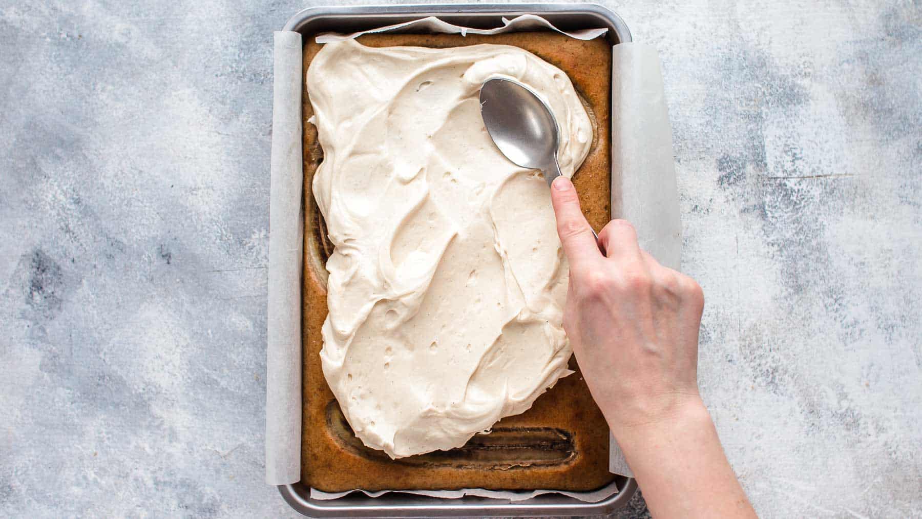 Spreading cream cheese frosting with back of a spoon on top of baked banana cake