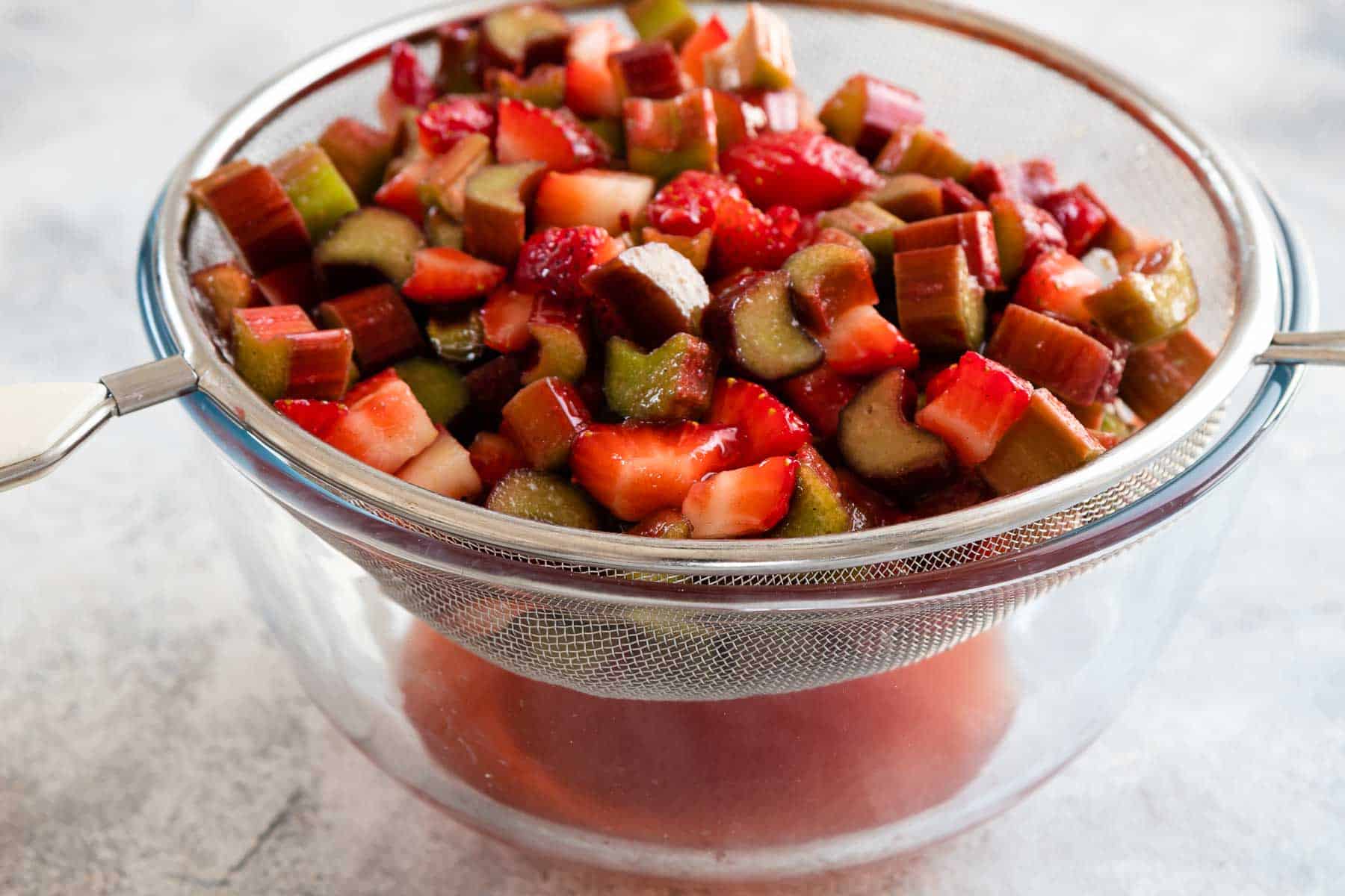 chopped and sugared strawberries and rhubarb in a mesh strainer placed on top of a bowl