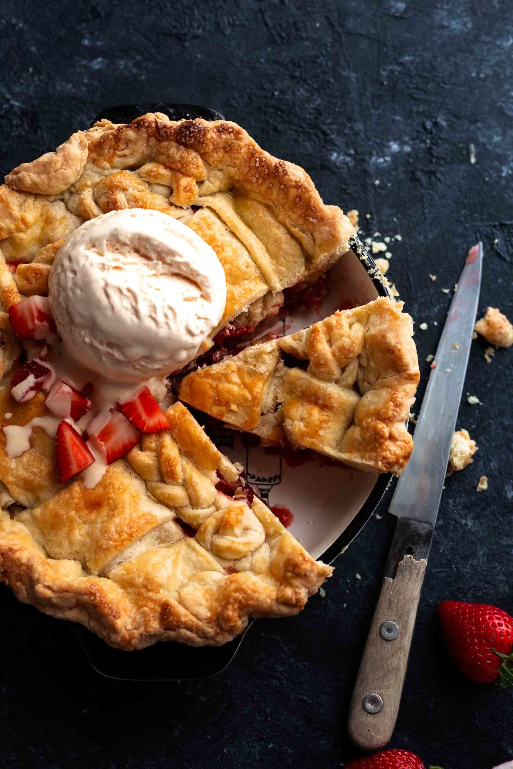 a sliced strawberry rhubarb pie in a baking dish with ice cream dollop on it and a knife next to it