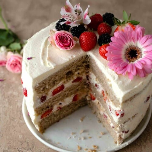 strawberry layer cake decorated with flowers and fruit on a cake plate with a quarter missing