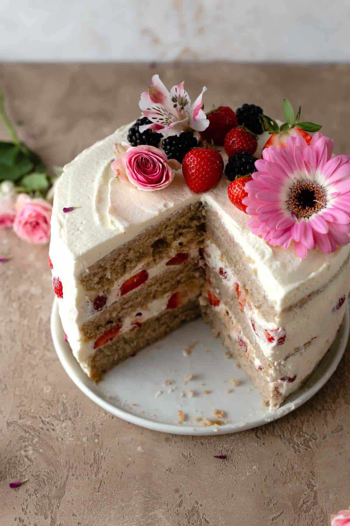 strawberry layer cake decorated with flowers and fruit on a cake plate with a quarter missing