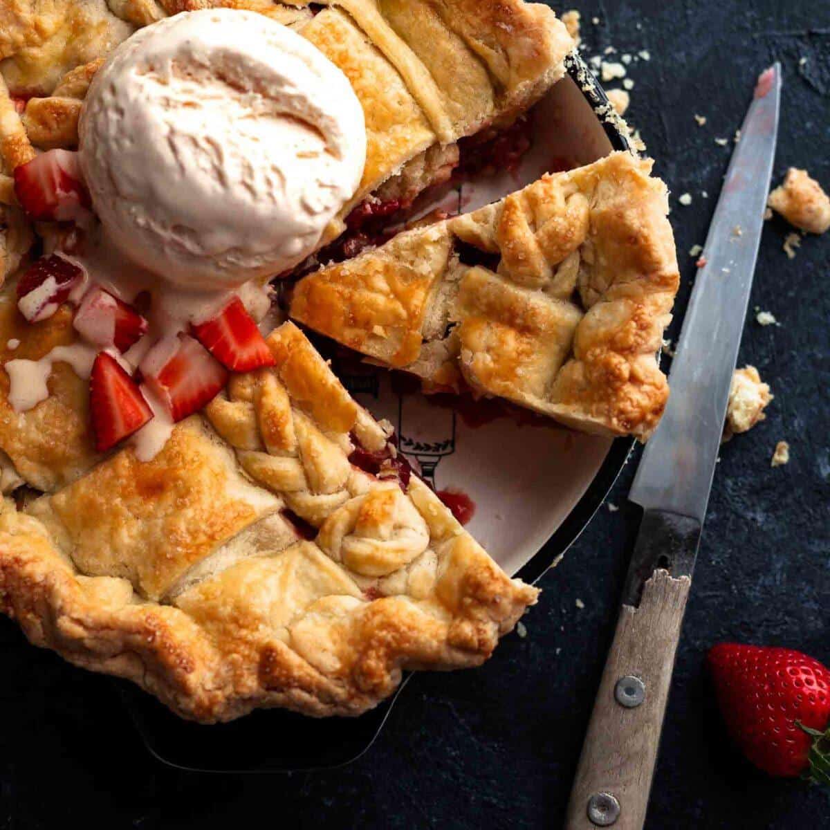 a sliced strawberry rhubarb pie in a baking dish with ice cream dollop on it and a knife next to it
