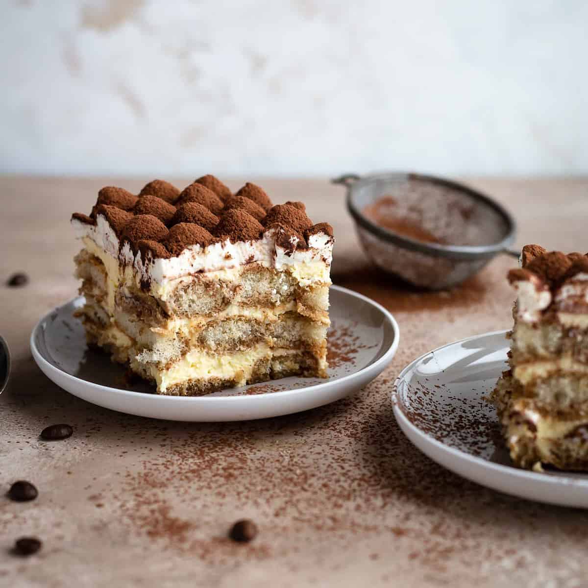 Two slices of tiramisu on two plates with a whipped cream and cocoa decoration on top