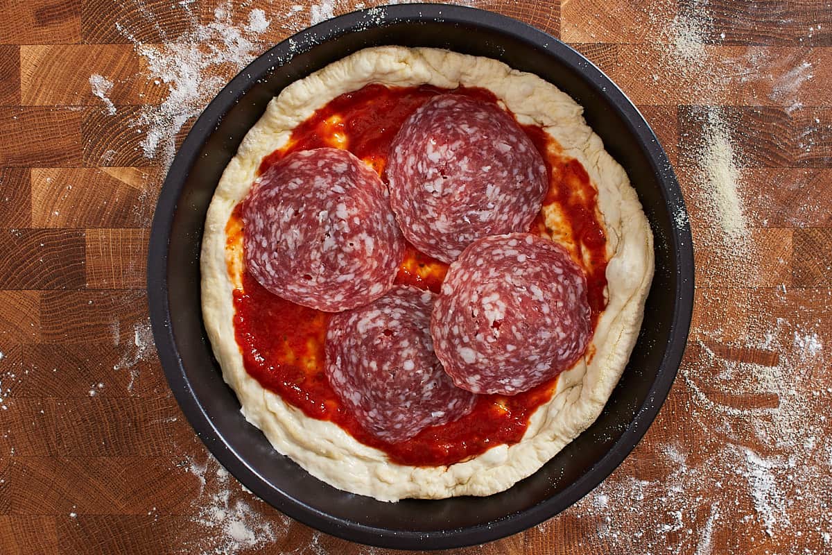 Pizza dough in a pizza pan topped with tomato sauce and pepperoni