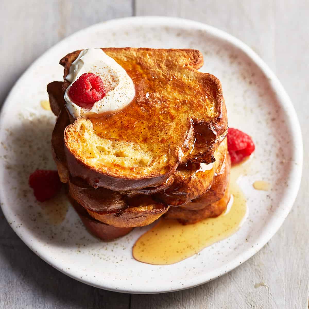 A stack of French toast topped with maple syrup, whipped cream, and raspberries