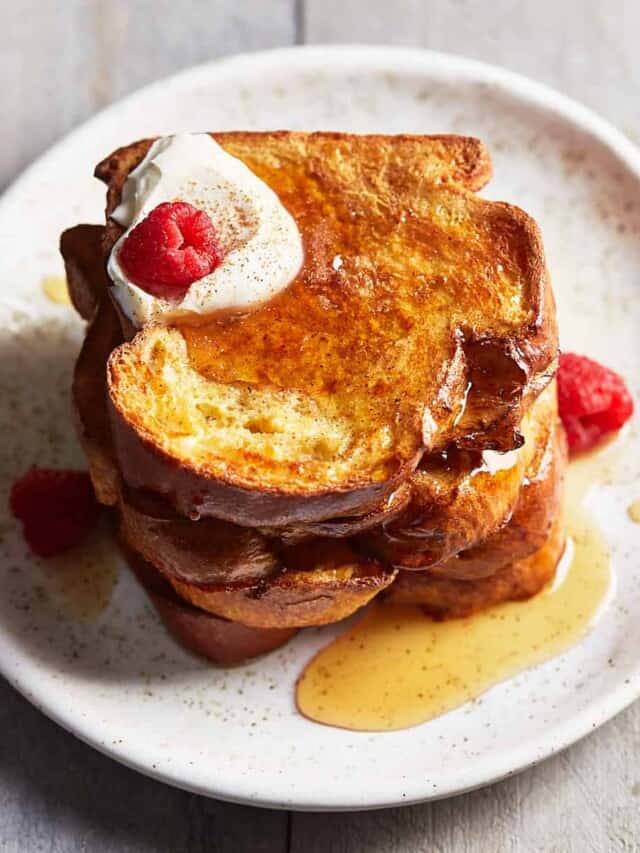 A stack of French toast topped with maple syrup, whipped cream, and raspberries