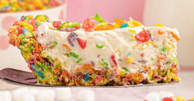 a slice of fruity pebbles cheesecake on a spatula on a colourful, pink background