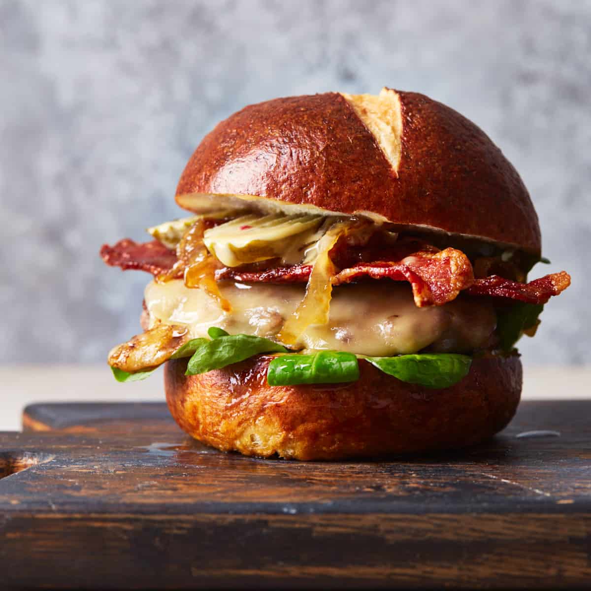 Pretzel bun filled with burger, cheese, bacon, sauce, lettuce, and pickles on a cutting board