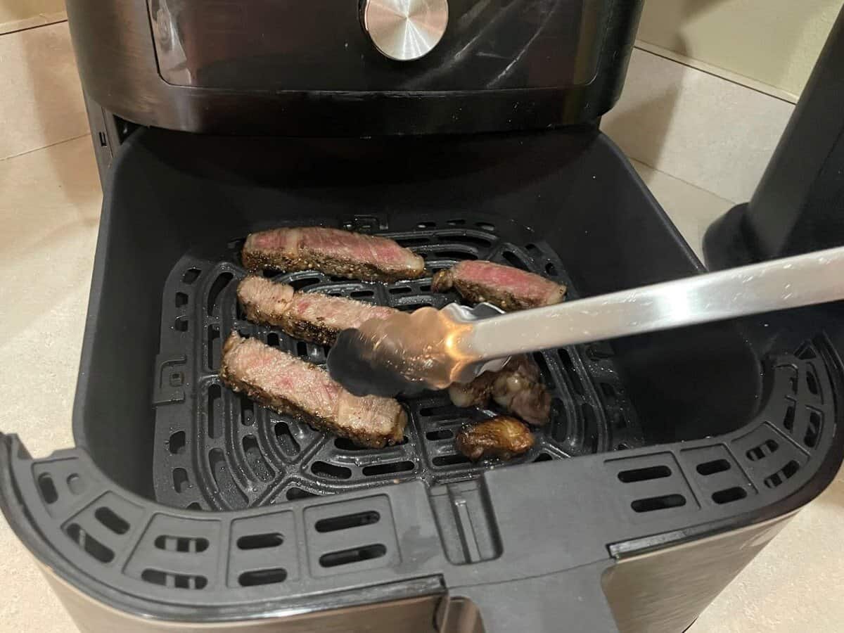 picking up steak slices with kitchen tongs in the air fryer basket