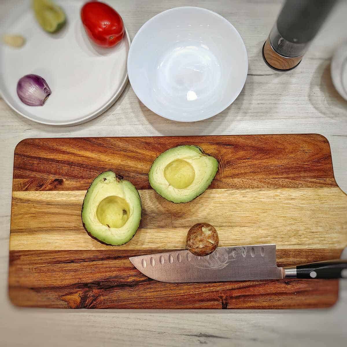 avocado on top of a cutting board with knife together with other ingredients in white bowls