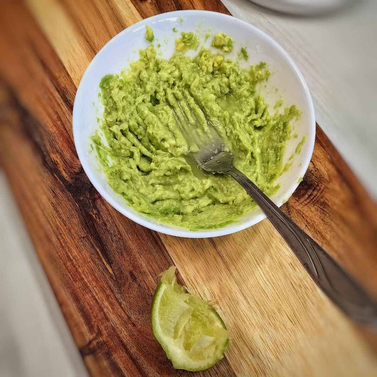 mashed avocado in a white bowl with a squeezed lime wedge on top of cutting board