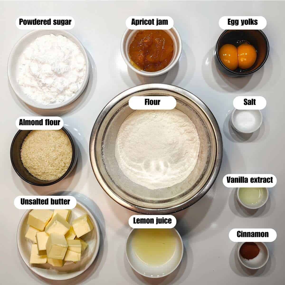 Ingredients to make linzer cookies in bowls on the table with text labels identifying them.