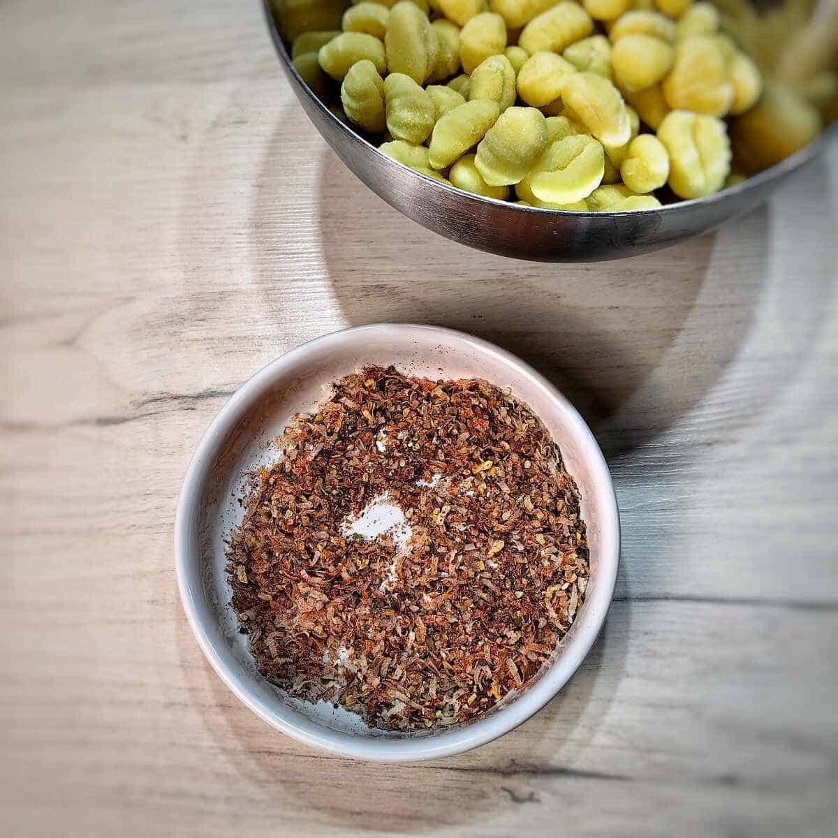 gnocchi in a mixing bowl and a white bowl with a mix of herbs and seasonings