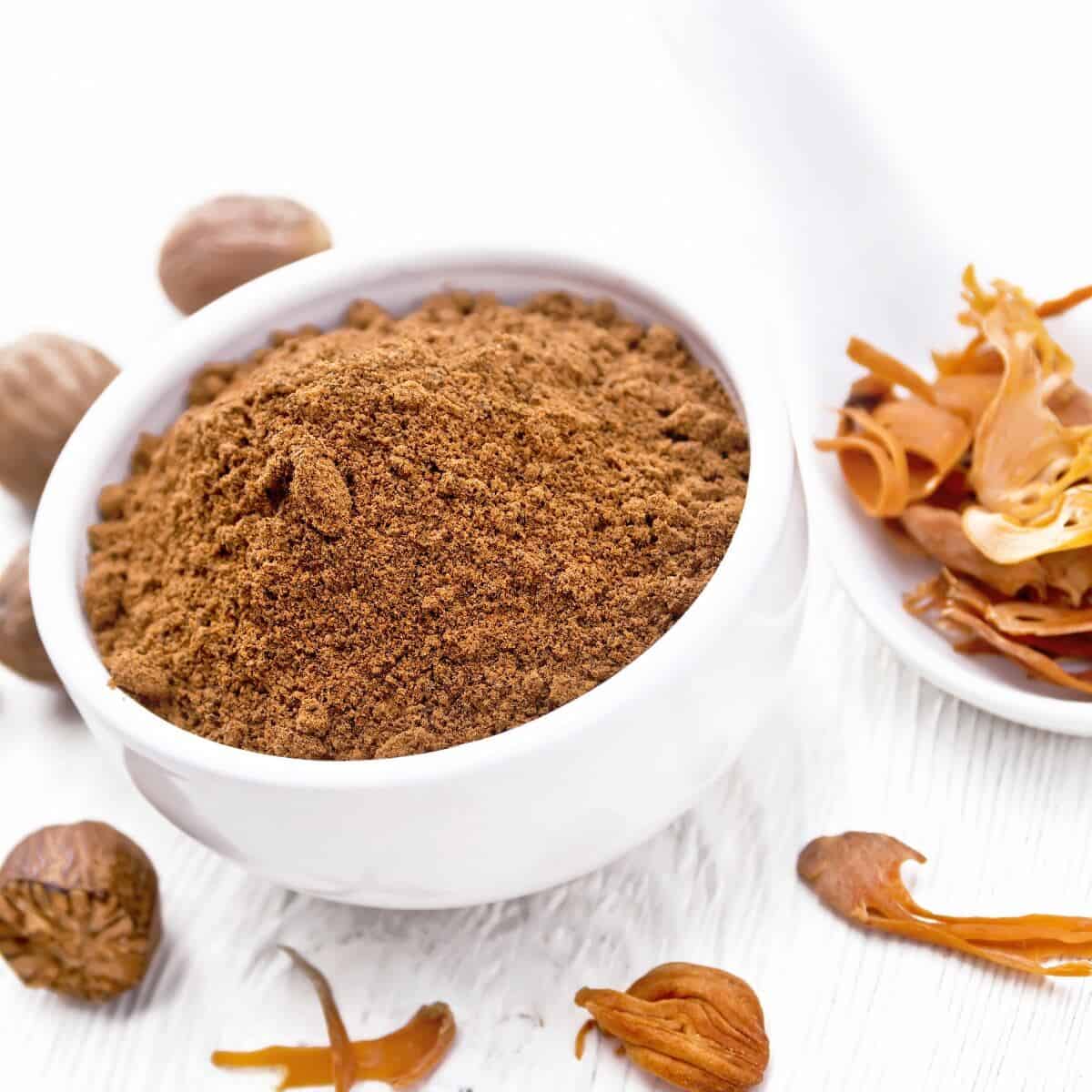 ground nutmeg in a bowl and dried nutmeg arillus in a spoon, whole nuts on white wooden board background