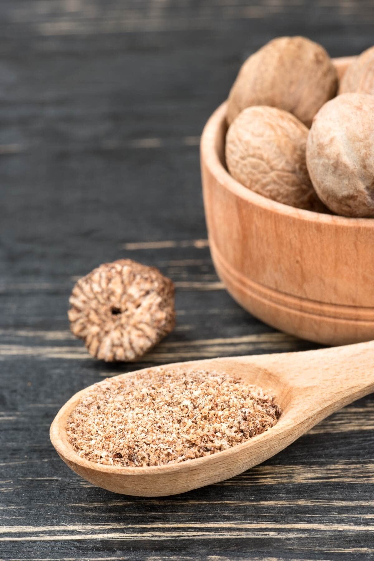 nutmeg powder in a spoon with a bowl of nuts on wooden background closeup