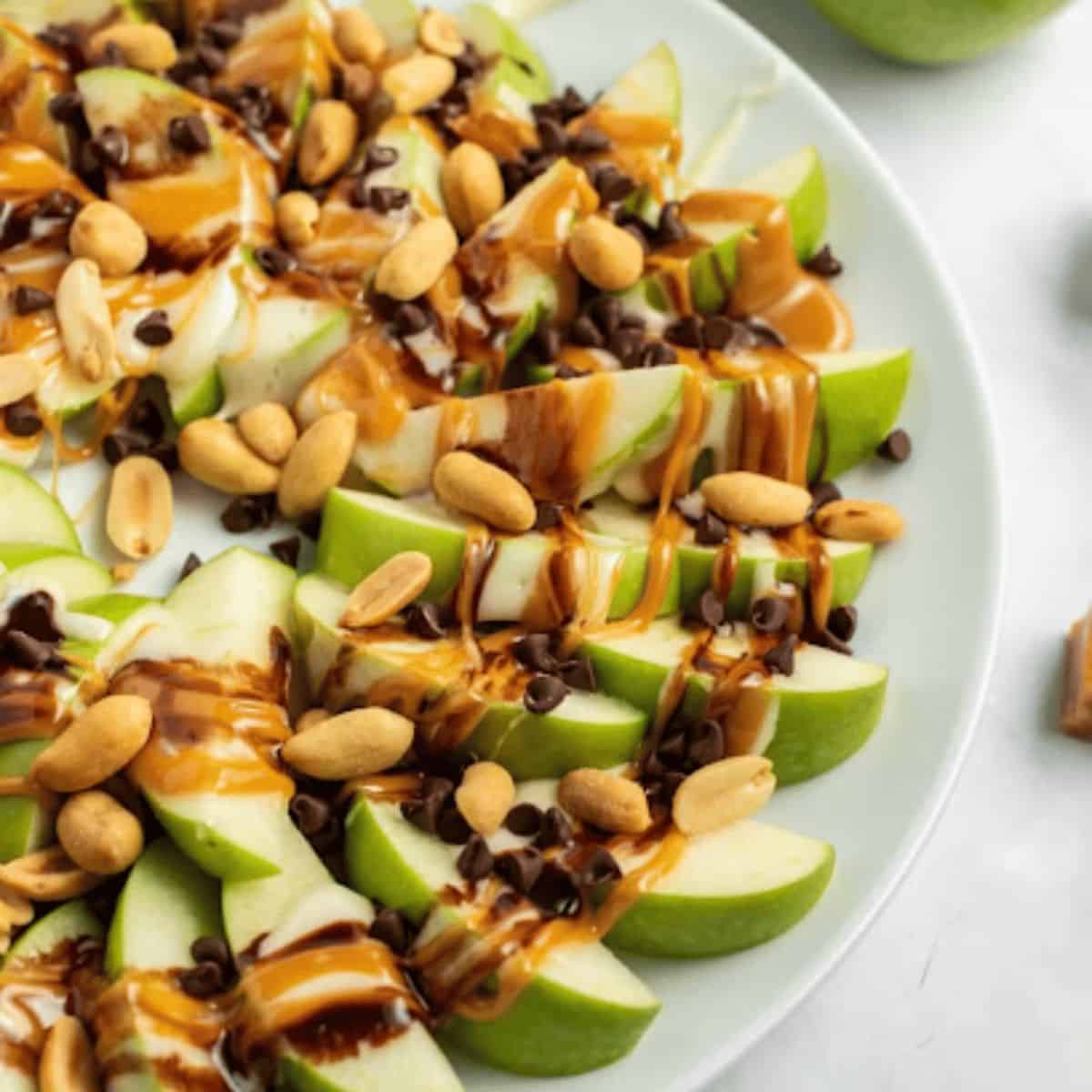 A colorful plate of apple nachos, featuring sliced apples drizzled with creamy peanut butter and melted caramel, topped with peanuts and mini chocolate chips.
