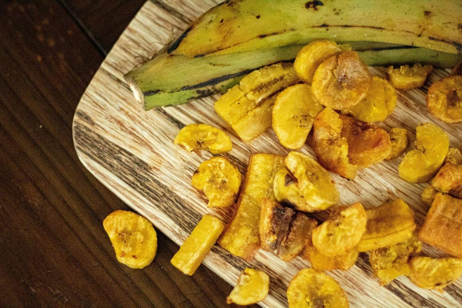 Two bananas that have not been peeled and bananas that have been sliced into different shapes on a wooden board.