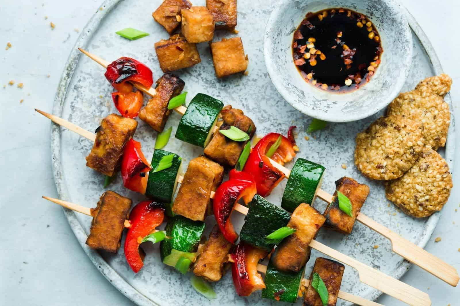 Tofu and various veggies on a stick alongside dipping sauce.