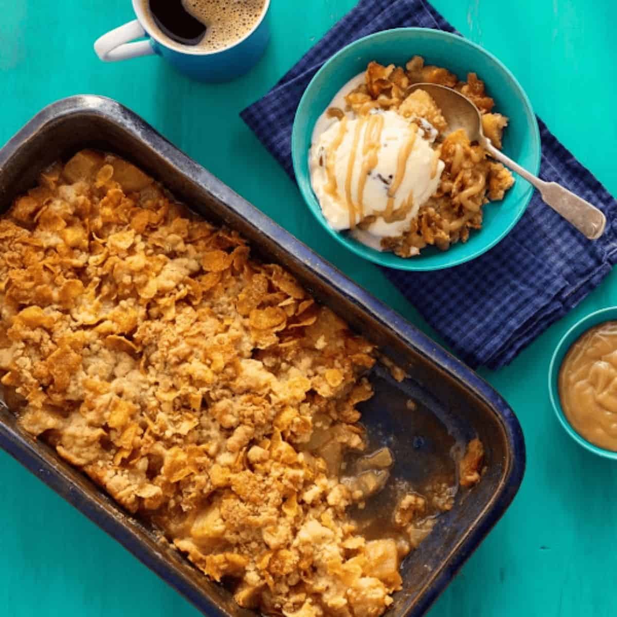 A warm butterscotch-cornflake apple crisp in a tray, featuring a golden-brown topping made of crunchy cornflakes and drizzled with butterscotch sauce.