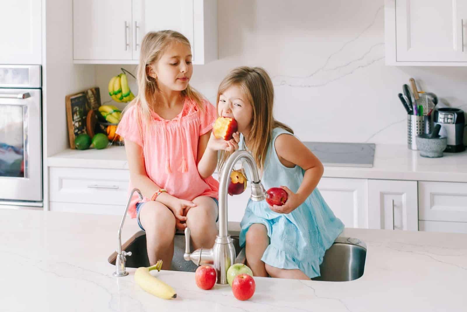 Two young girls sitting on a counter eating apples over the sink. 