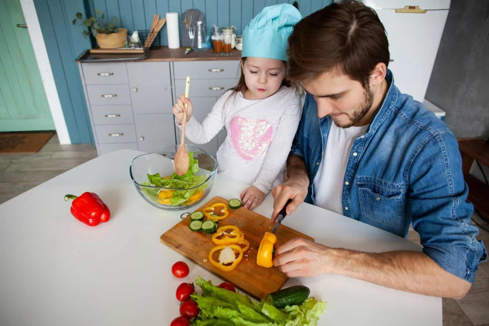A father cutting peppers with a knife while his daughter is stirring ingredients with a wooden spoon in a clear dish