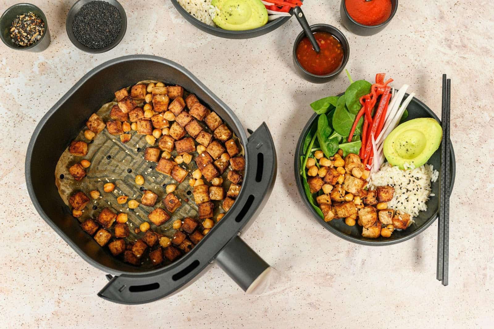 An air fryer basket with bites in it and a bowl on the side with bites, avocado, rice, fresh herbs, and chopsticks.