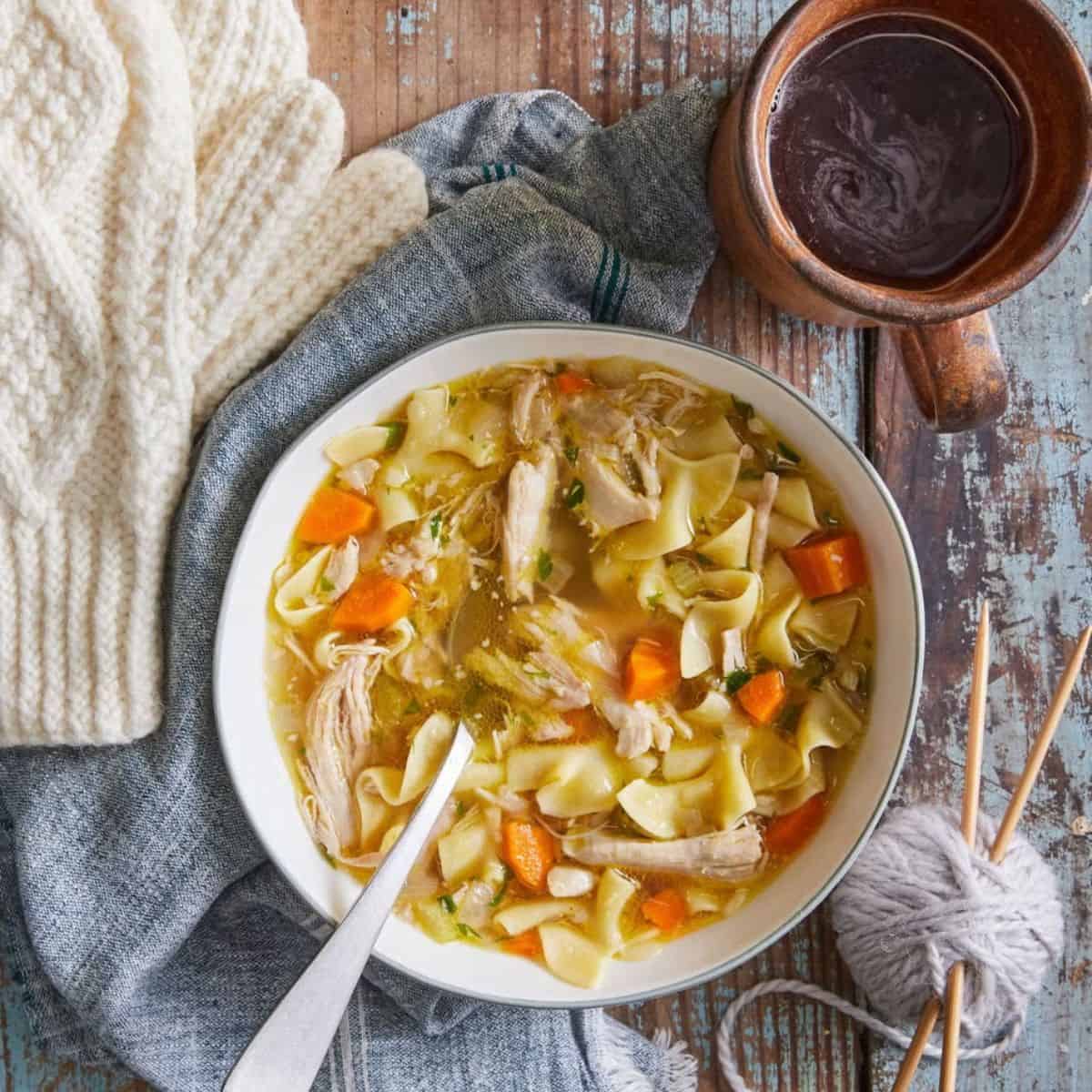 Delicious chicken noodle soup in a circular bowl with a metal spoon, and a cup of coffee on the table. 