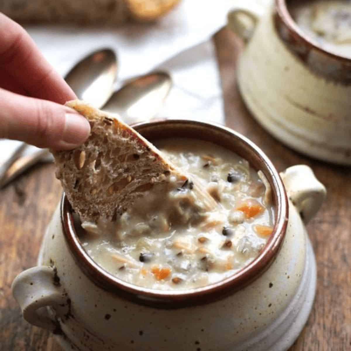 A yummy treat with slow cooker wild rice soup and a person dunking a piece of whole wheat bread in it.