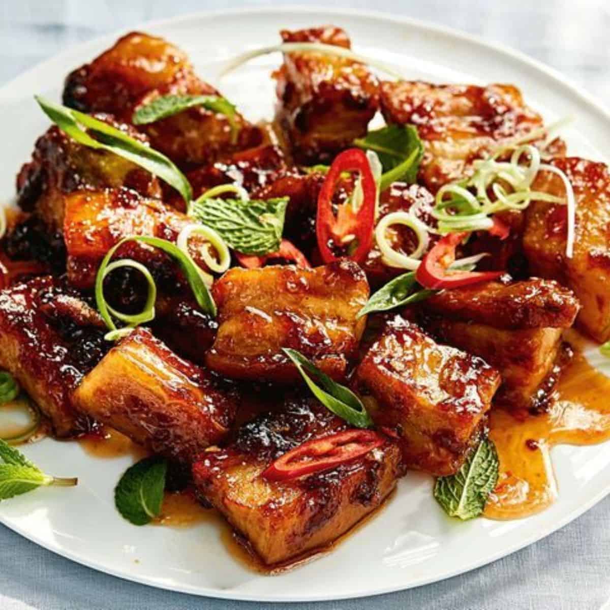 Juicy pork belly bites topped with veggies, served in a white plate. 