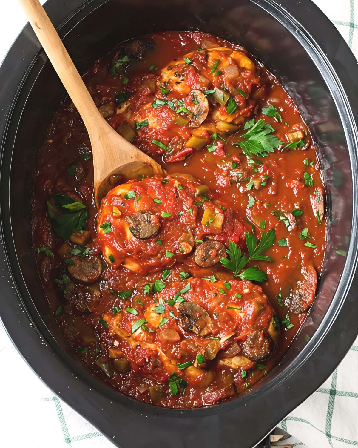 Healthy crockpot chicken cacciatore with tomatoes, mushrooms, and fresh parsley in a slow cooker with a wooden spoon.