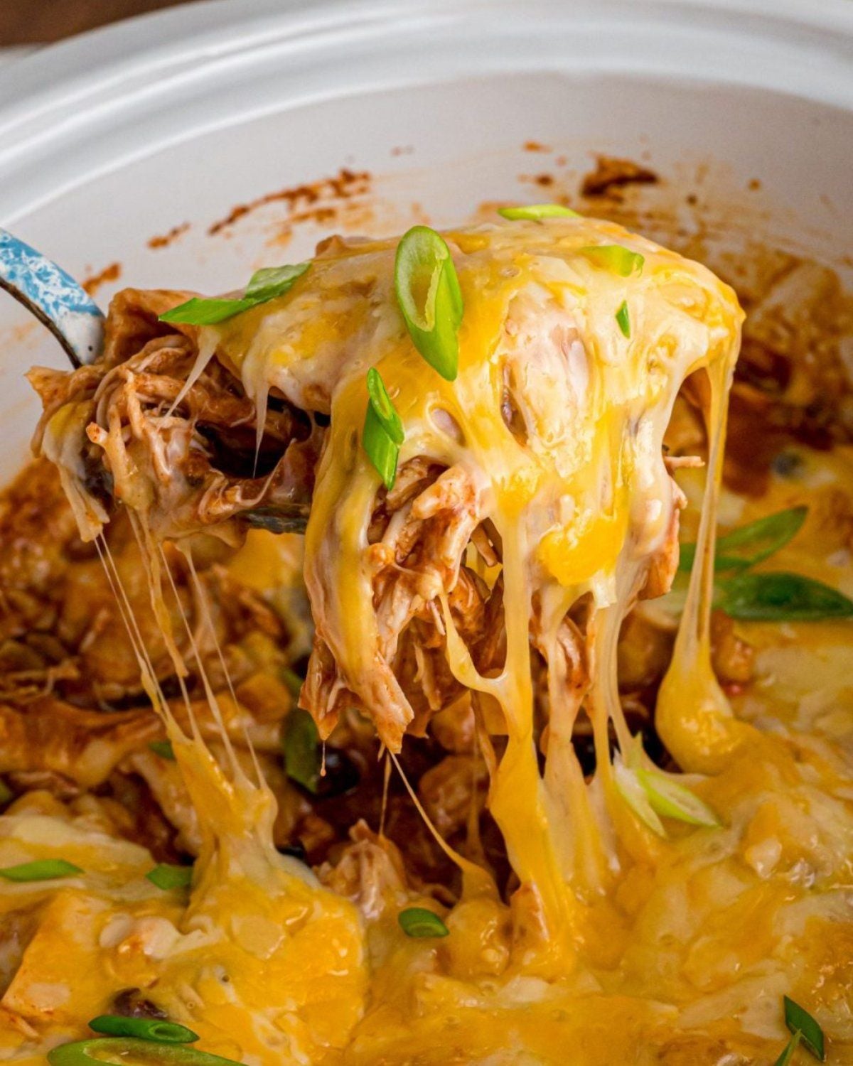 Slow cooker high protein enchilada casserole being scooped with melted cheese stretching from a crockpot garnished with green onions.