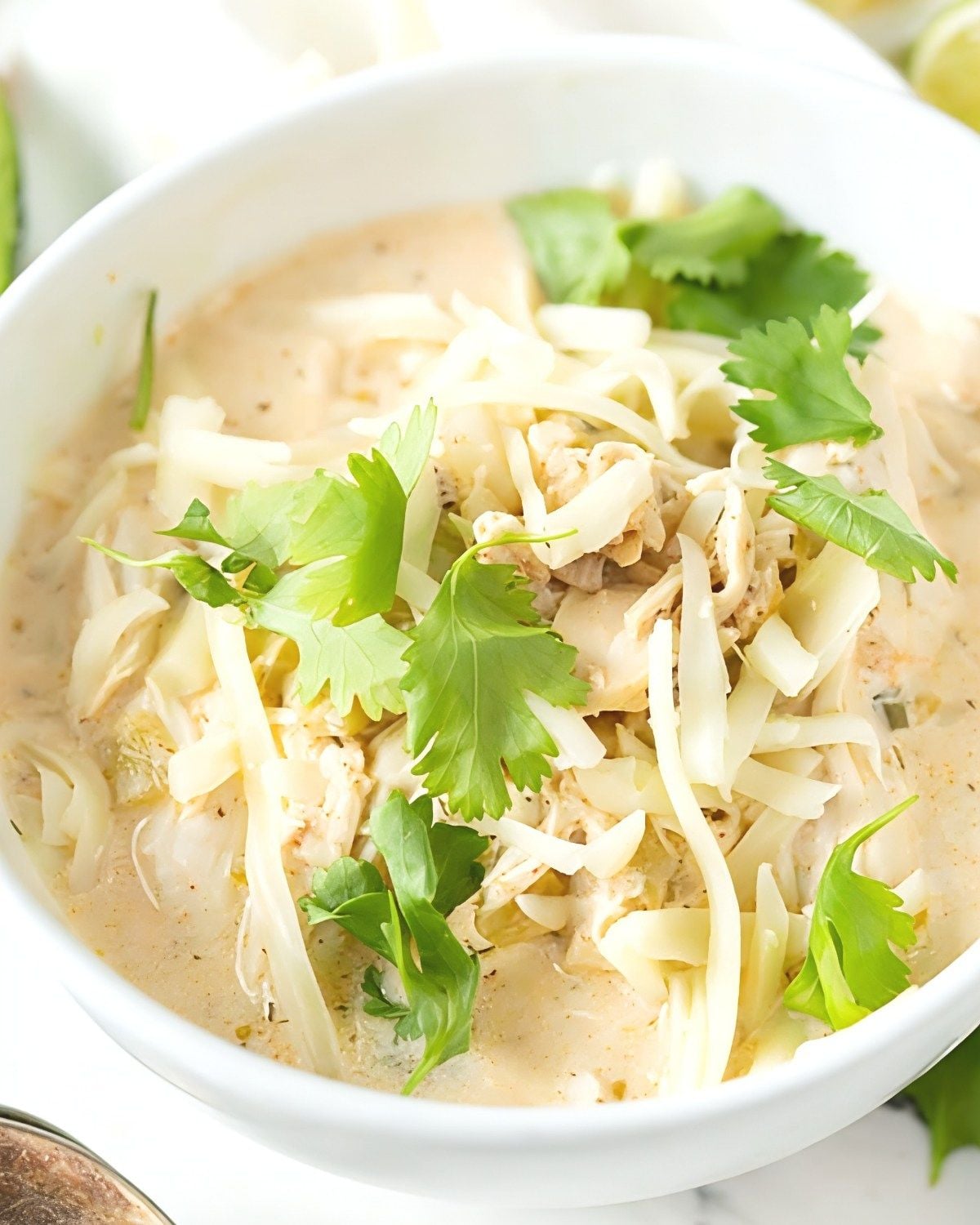 A white bowl filled with creamy white chicken chili, topped with shredded white cheese and fresh cilantro leaves