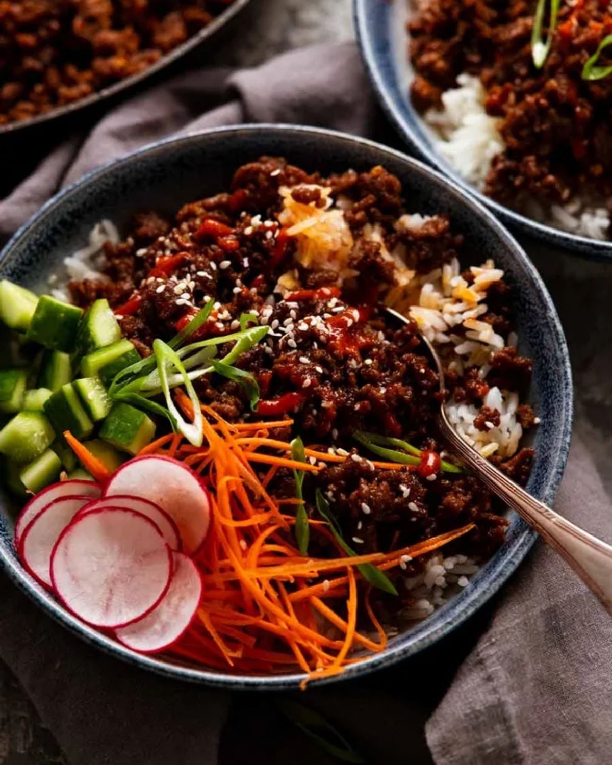 Ground meat crumbles served over white grains with sliced cucumbers, radishes, shredded carrots, and green onion garnishes inside a bowl