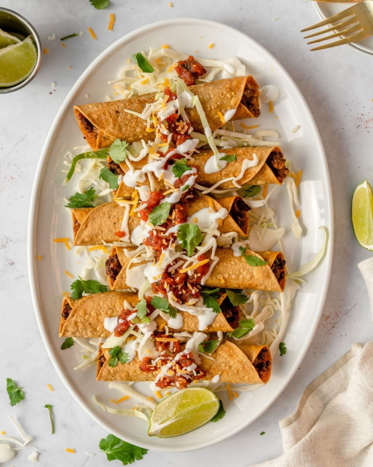 Crisp rolled corn tortillas filled with seasoned meat, drizzled with white crema, salsa, shredded cabbage, and cheese on a white platter.