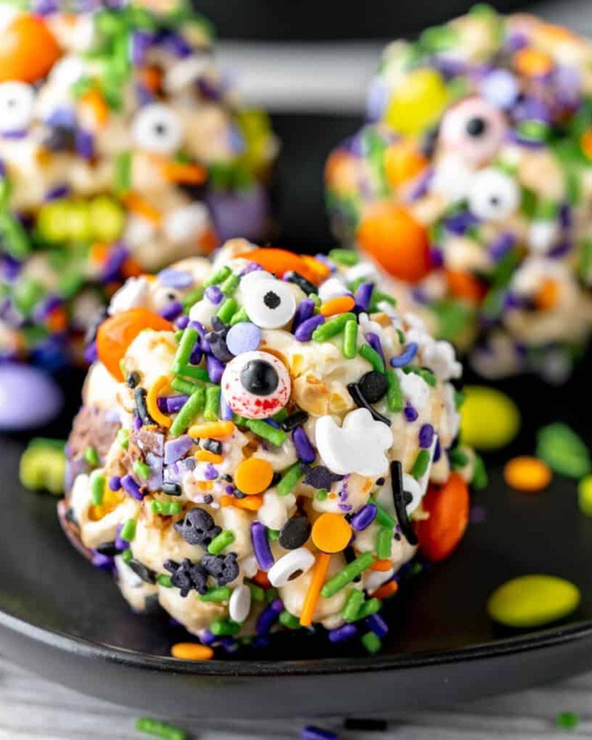 A close-up photograph captures a round popcorn ball heavily decorated with a festive Halloween mix of sprinkles and candies.