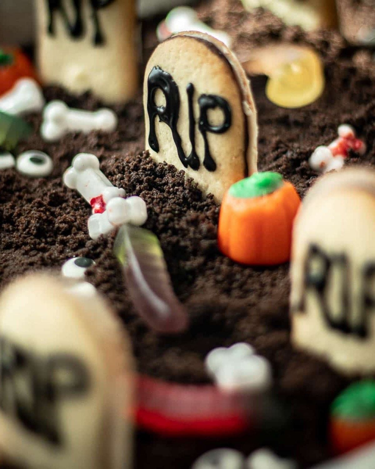 Here is concise alt text for your image:
Alt text: A close-up view of Halloween graveyard brownies decorated to look like a spooky cemetery. The dessert features crushed chocolate cookie crumbs acting as dirt, upright cookie tombstones with "RIP" written in black icing, small candy pumpkins, bone-shaped candies, and candy gummy worms scattered throughout