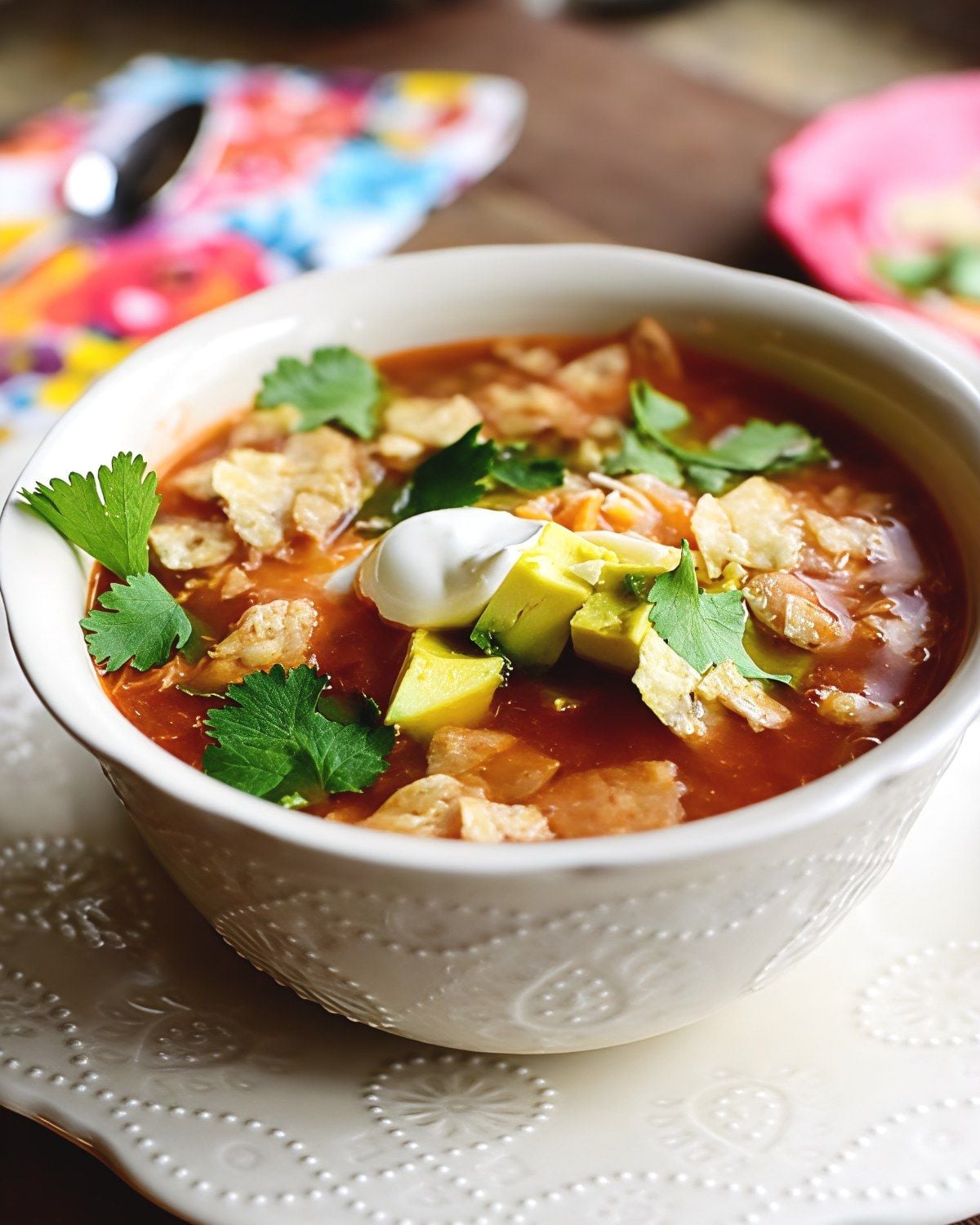 Healthy slow cooker chicken tortilla soup in a white patterned bowl, topped with diced avocado, a dollop of sour cream, crushed tortilla chips, and fresh cilantro.