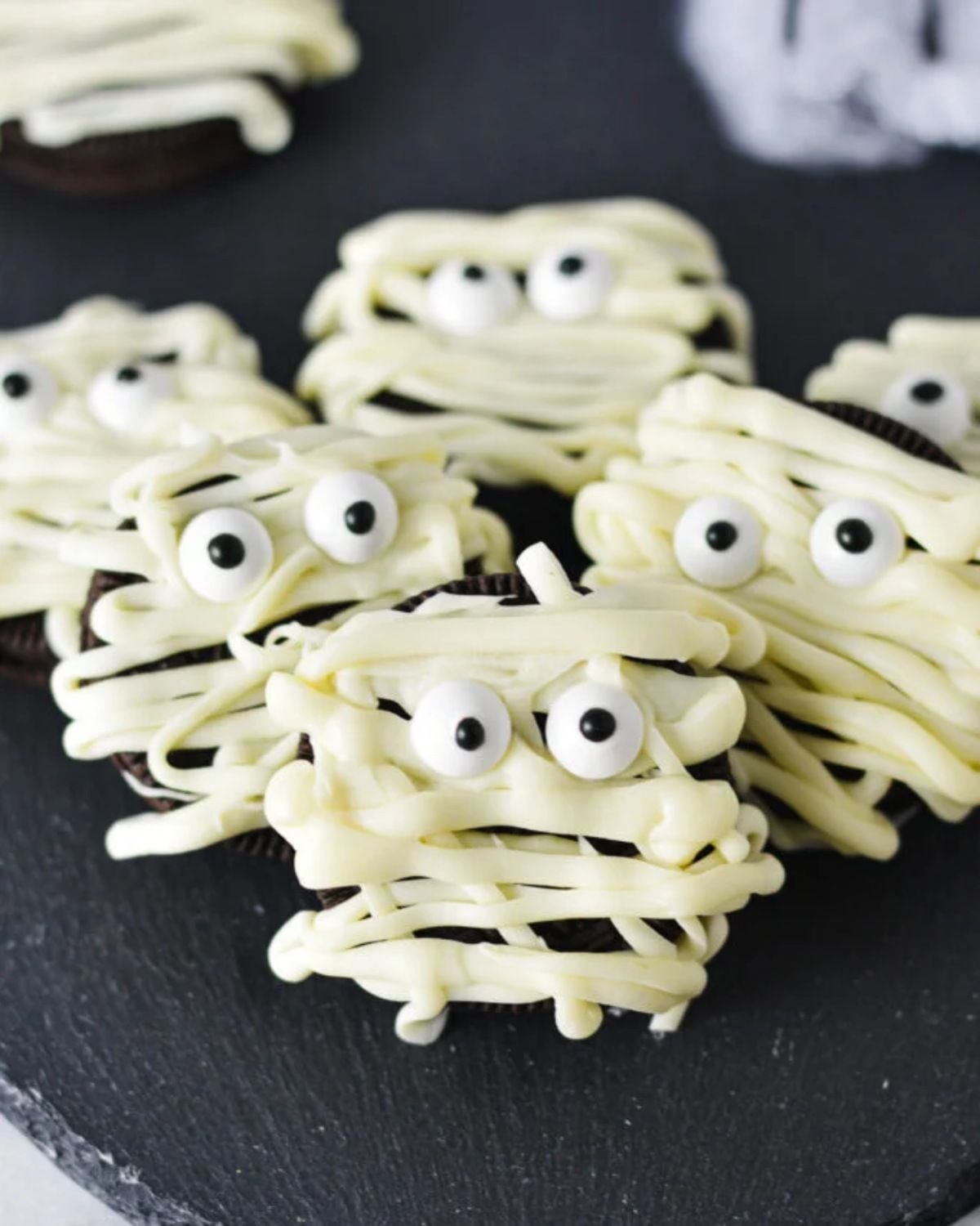 A close-up of several Oreo mummies arranged on a dark plate, decorated to look like spooky Halloween treats. Each cookie is drizzled with white chocolate to resemble mummy bandages and features two small edible candy eyes peeking through