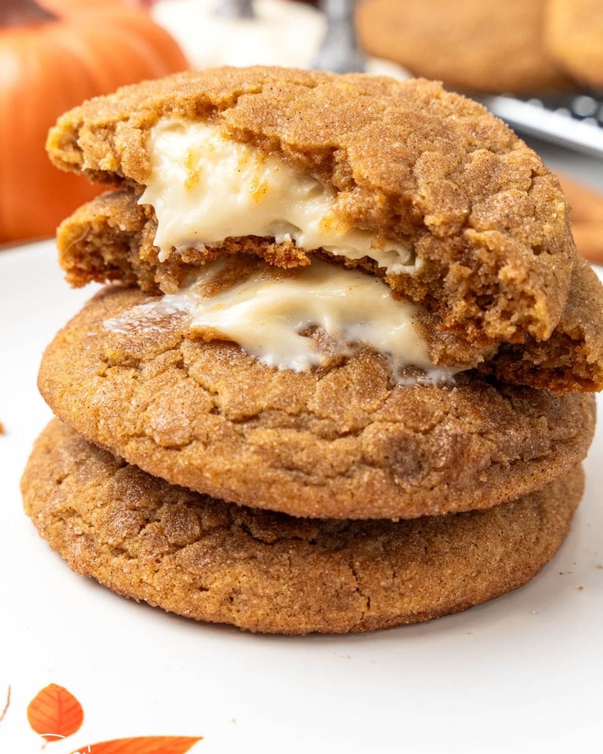A stack of three soft-baked pumpkin cheesecake cookies on a white plate, with the top cookie broken in half to reveal a creamy, melted cheesecake filling inside. A small pumpkin is visible in the upper-left background.