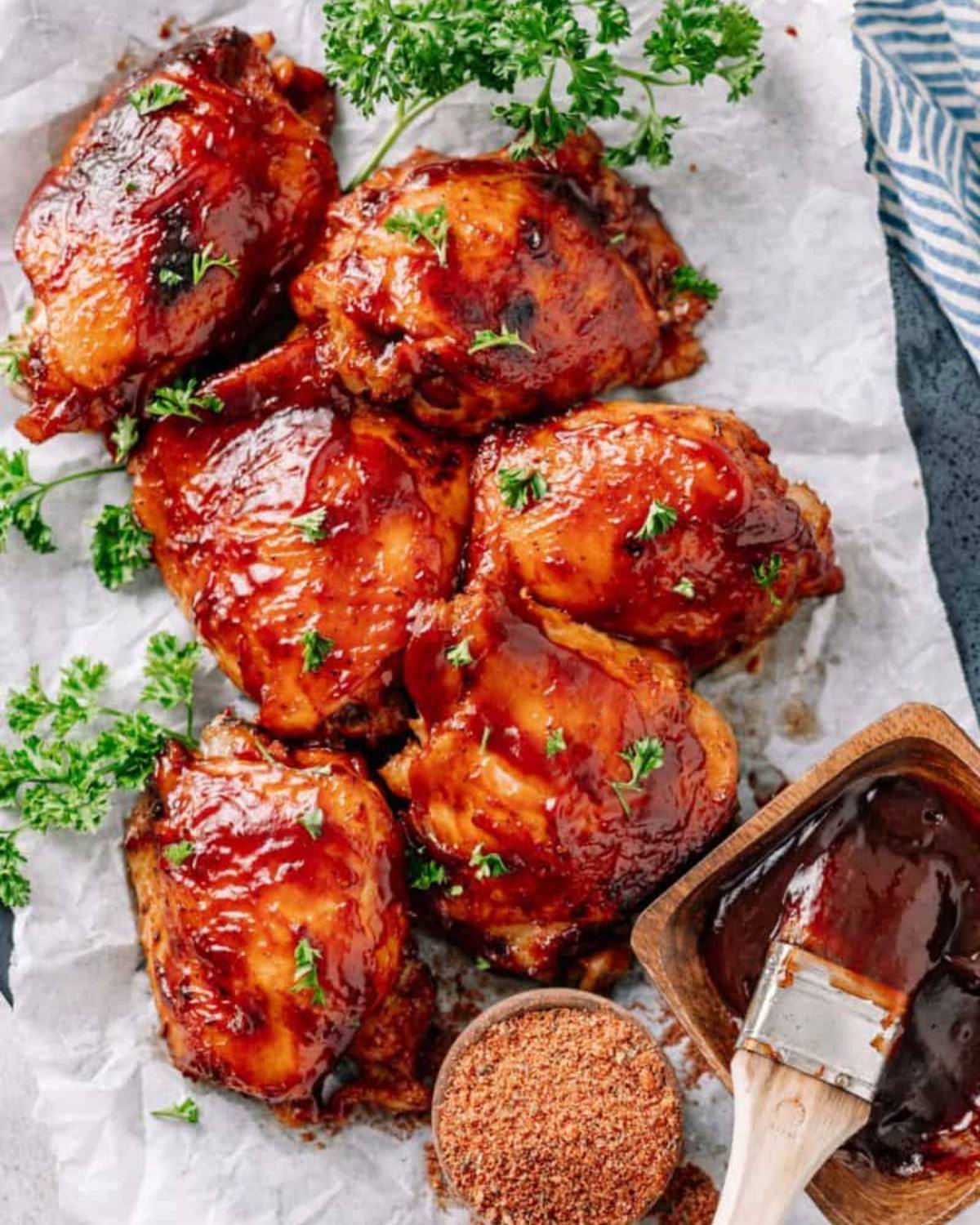 Slow cooker high protein BBQ chicken thighs garnished with parsley on parchment paper next to a small wooden bowl of spice rub and a basting brush with barbecue sauce