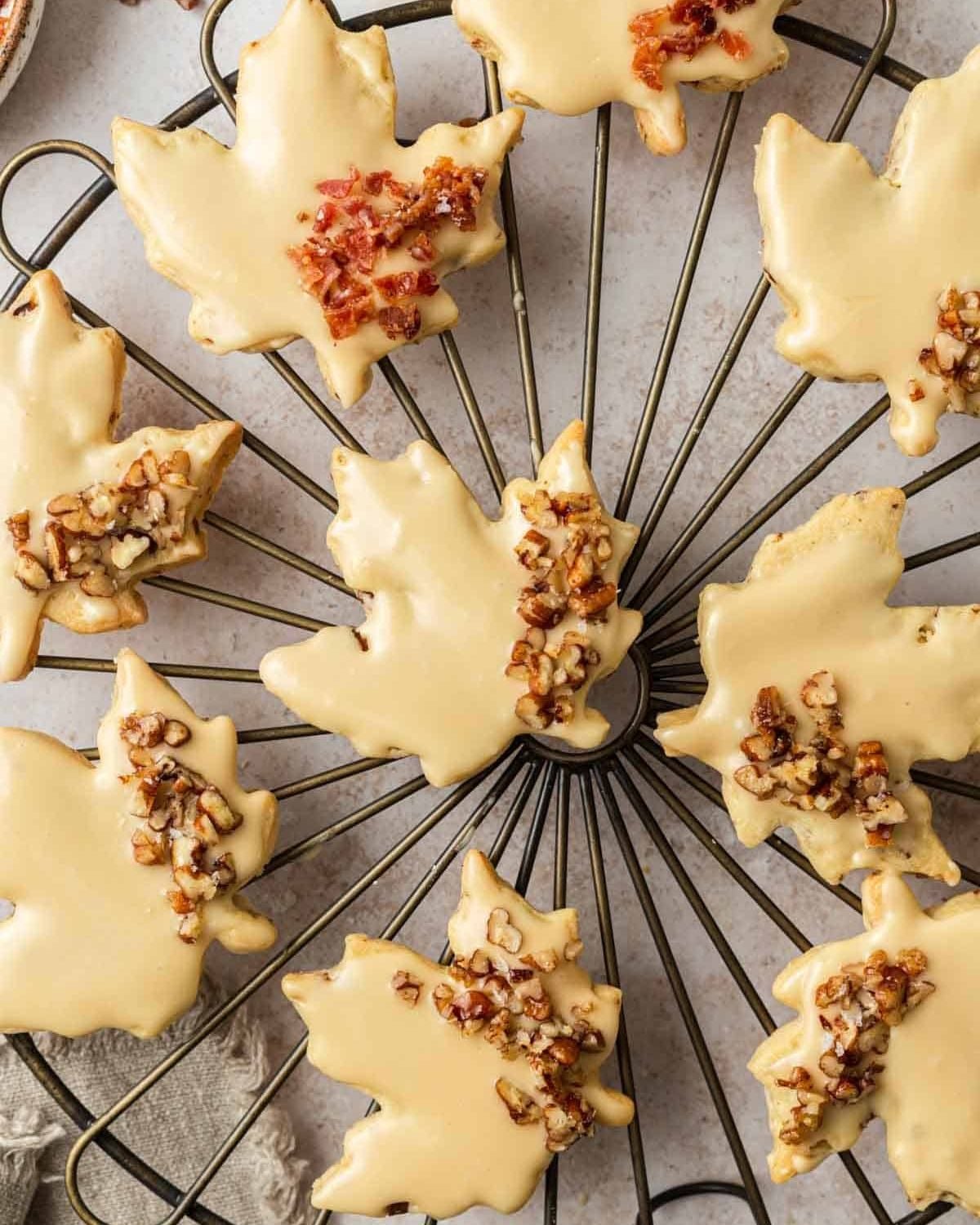 Maple leaf-shaped shortbread cookies glazed with a smooth icing and garnished with chopped pecans, arranged on a wire cooling rack.