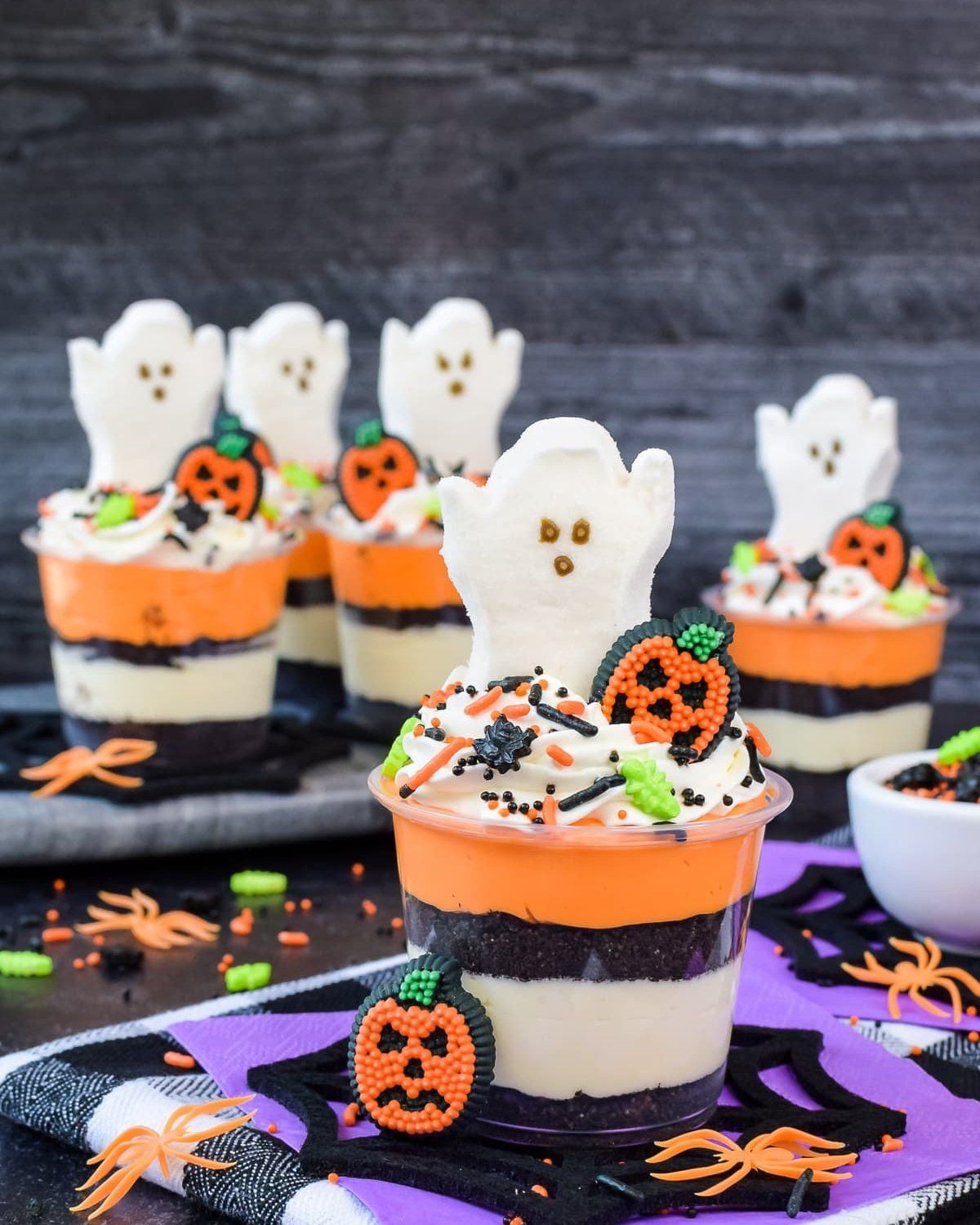Layered Halloween cheesecake cups in plastic cups topped with a white ghost cookie, a pumpkin candy, and Halloween sprinkles, set against a dark rustic background with spider web decor.