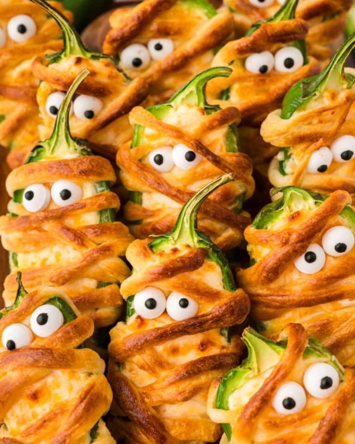 A tray of spooky jalapeno popper mummies wrapped in dough with candy eyes, perfect for a Halloween party snack.