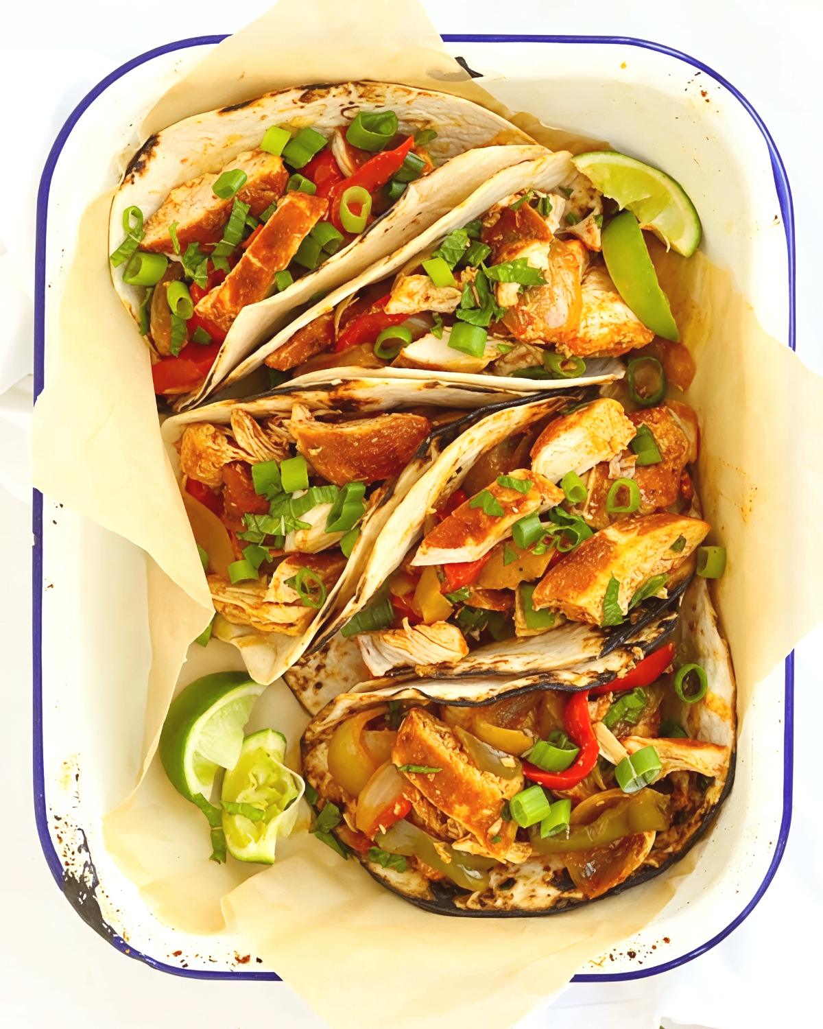 Healthy crockpot chicken fajitas filled with bell peppers, onions, and topped with green onions in a baking dish with lime wedges.