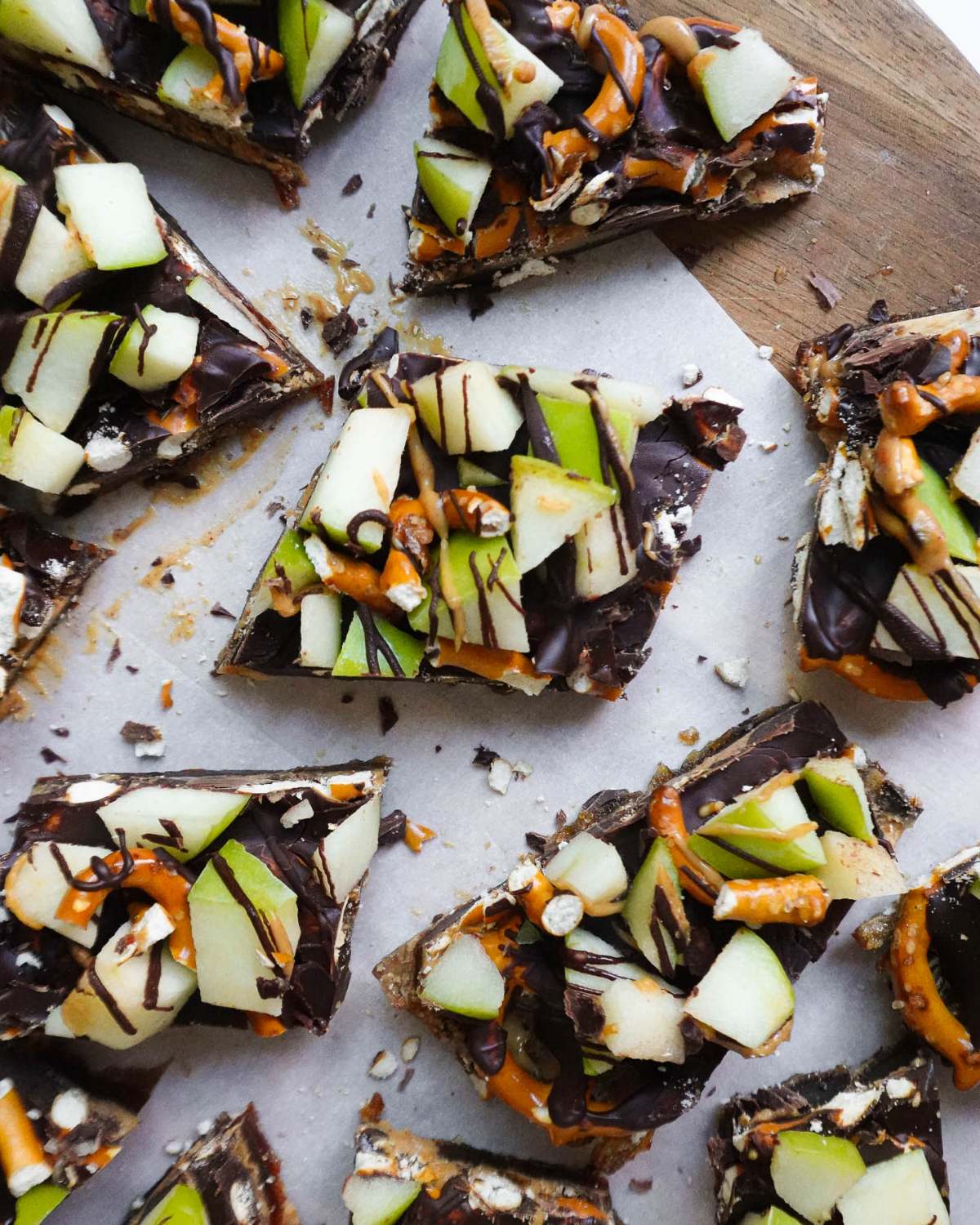 Healthy fall dessert chocolate apple bark pieces topped with green apples, pretzels, and drizzles on parchment paper and a wooden board